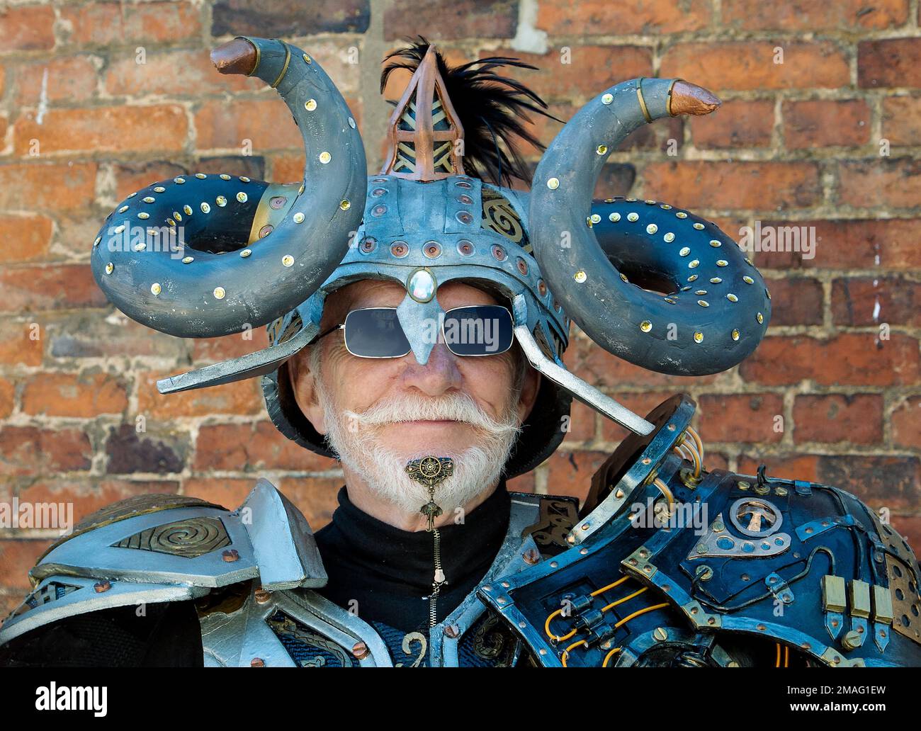 Ein exzentrisch aussehender Mann mit weißem Bart, mit futuristischer Rüstung im Retro-Look und einem gewellten Hornhelm. Älterer, männlicher Steampunk mittleren Alters. Stockfoto