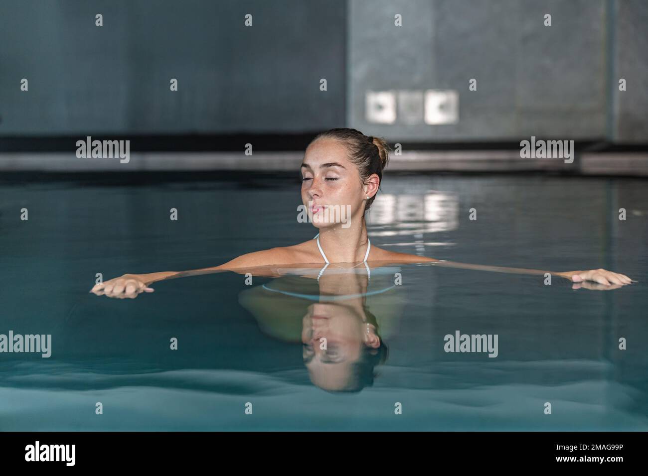 Eine ruhige junge Frau mit einem Haarbrötchen, das die Augen schließt und während der Spa-Sitzung im Resort im klaren Wasser im Pool schwimmt Stockfoto