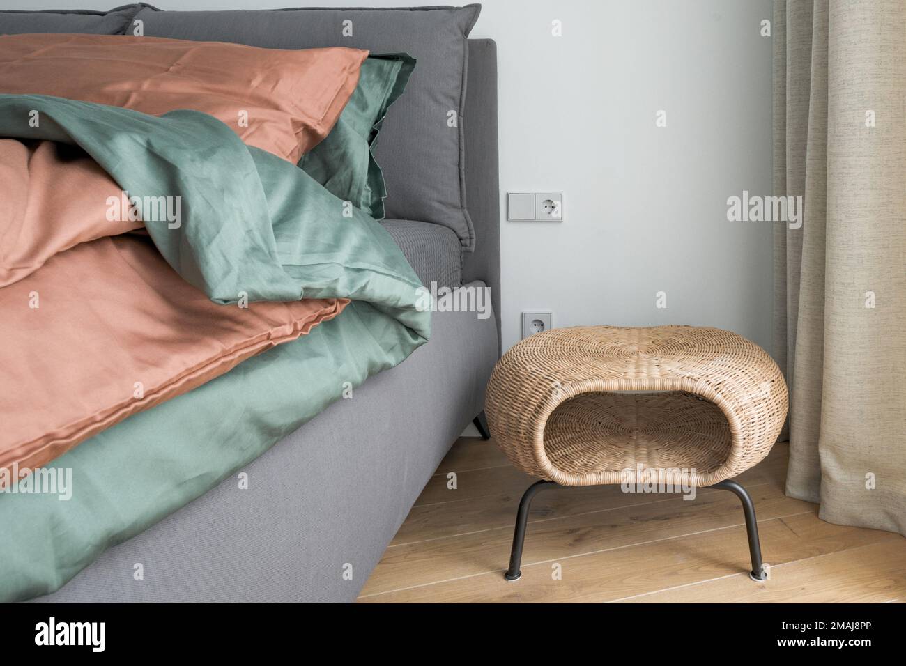 Zimmereinrichtung mit rostfarbener Bettwäsche und Kissen auf einem Bett, grauer Decke, Rattan Nachttisch und Wandleuchte aus Metall. Stockfoto