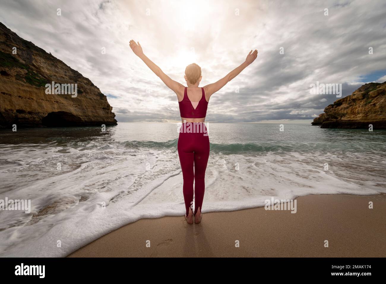 Rückansicht einer sportlichen Frau, die am Meer steht, mit erhobenen Armen, um dem Alltag zu entfliehen. Stockfoto