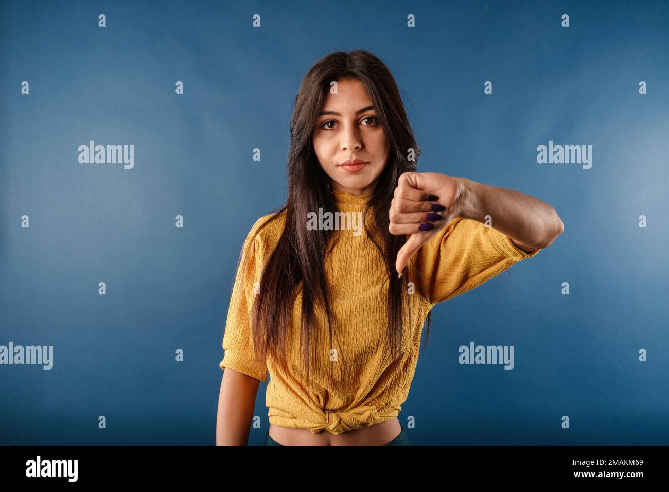 Das Porträt einer jungen brünetten Frau, isoliert auf blauem Hintergrund, macht ein Daumen-nach-unten-Schild, während sie mit einem unzufriedenen Ausdruck in die Kamera schaut. Nein. Nein Stockfoto