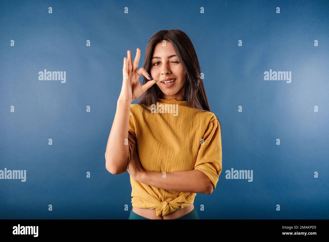 Junge dunkelhaarige Frau, die ein gelbes Oberteil trägt, isoliert auf blauem Hintergrund, stimmt Ihnen vollkommen zu, sie zeigt gute Zeichen- und Nicken-Zustimmung. Blinzend und immer Stockfoto