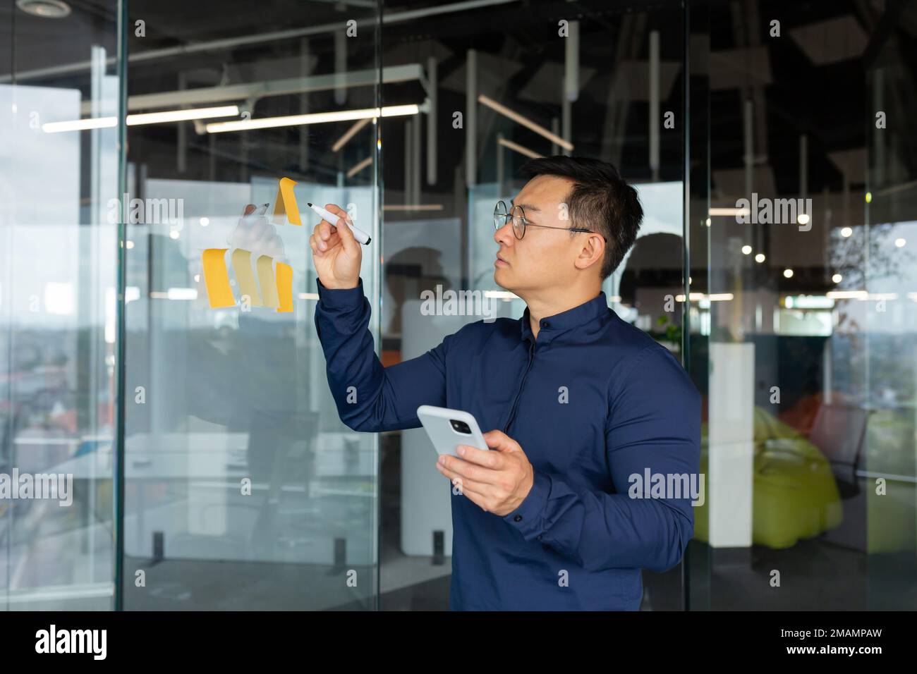 Asiatischer Mann in Gedanken, der Pläne und Strategien auf farbigem Papier auf einer Glaswand aufschreibt, Brainstorming bei Geschäftsleuten bei der Arbeit. Stockfoto