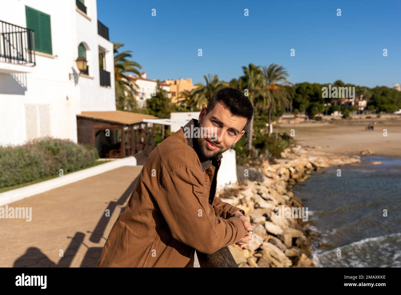 Junger Mann mit positivem Bart in brauner Jacke, der sich auf den Zaun lehnt, während er neben dem Meer und exotischen Bäumen unter wolkenlosem blauem Himmel steht und in die Kamera schaut Stockfoto
