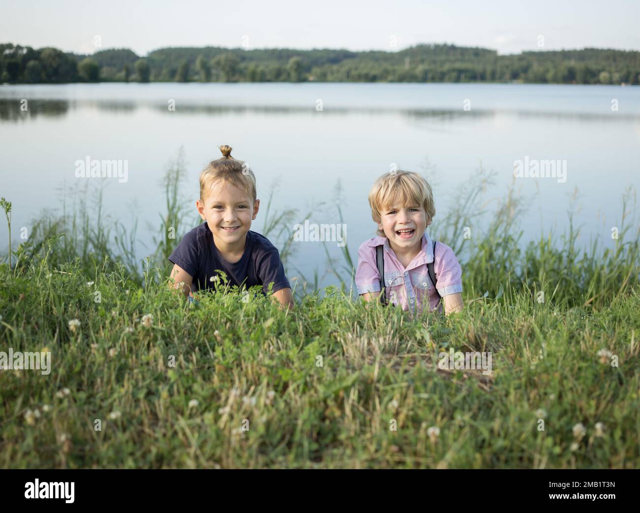 Zwei glückliche Kinder liegen auf dem Bauch im Gras in der Nähe des Sees. Lächelnde Freunde, Jungs, ruhen sich in einem Sommerpark aus. Gesunder Lebensstil. Joy, positi Stockfoto