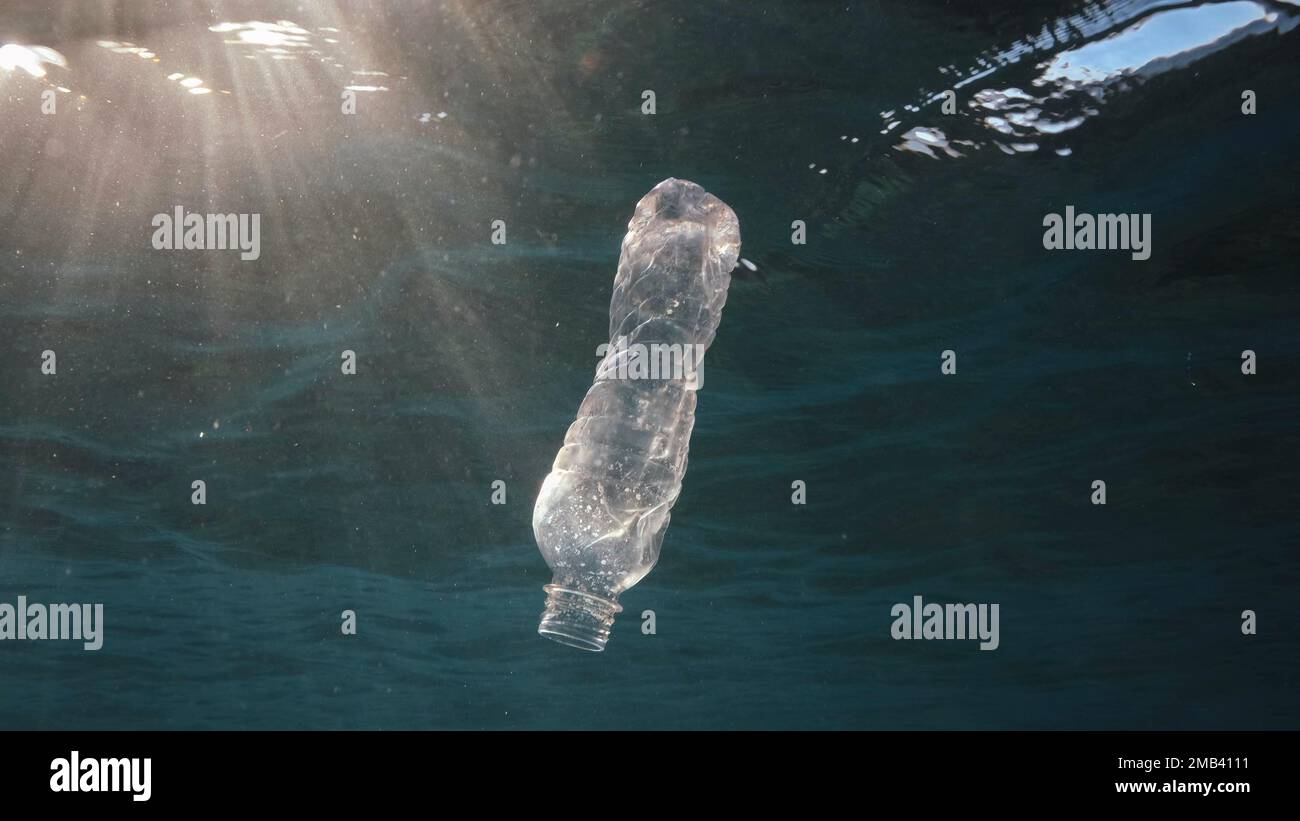 Plastikverschmutzung, weggeworfene Plastikflasche, die unter der Wasseroberfläche in Sonnenstrahlen am Sonnenuntergang driftet. Plastikflasche in blauem Wasser. Plastikmüll Stockfoto