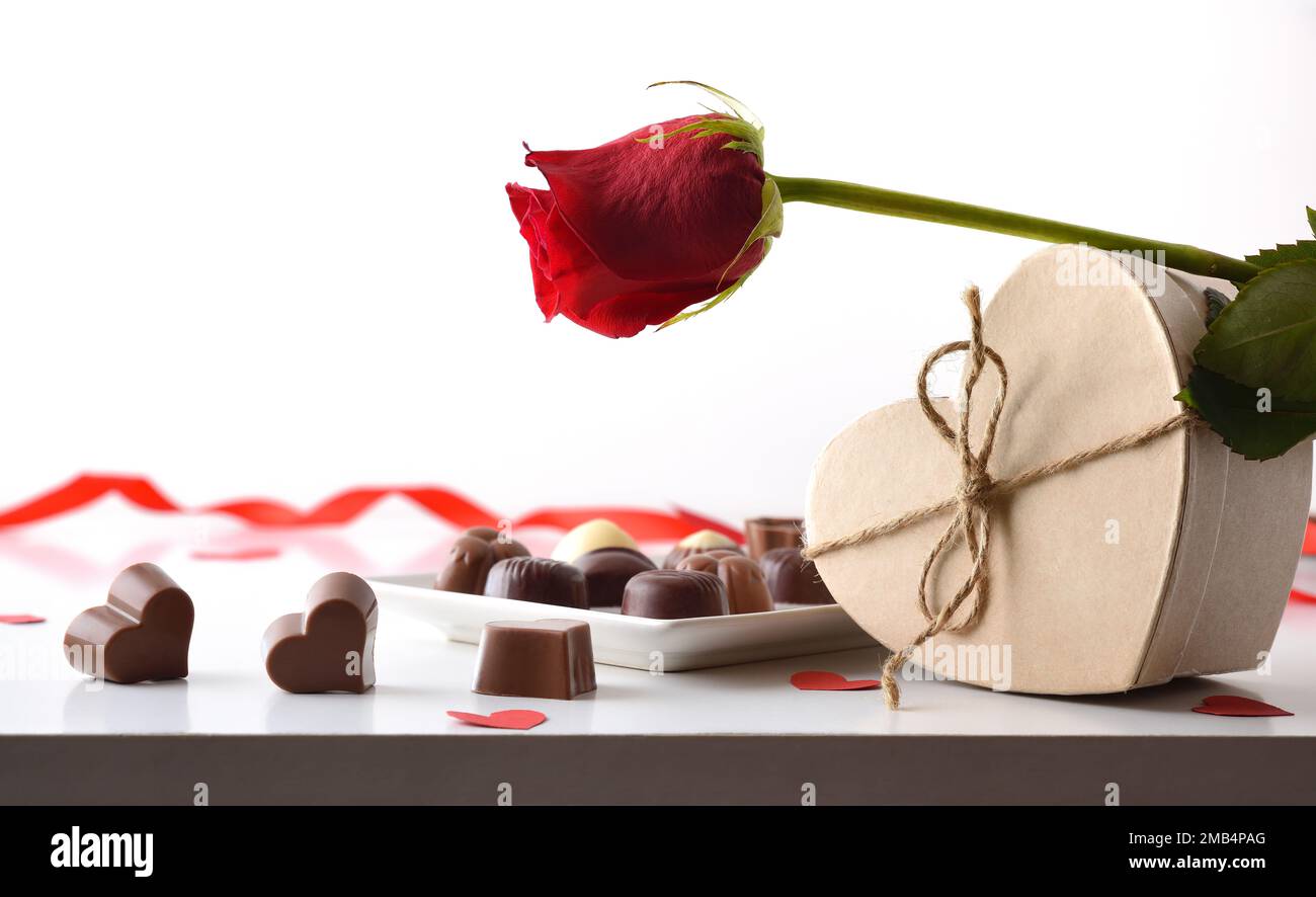 Auswahl an Pralinen auf weißem Tisch mit roter Rose auf herzförmiger Schachtel und isoliertem weißem Hintergrund. Vorderansicht. Stockfoto