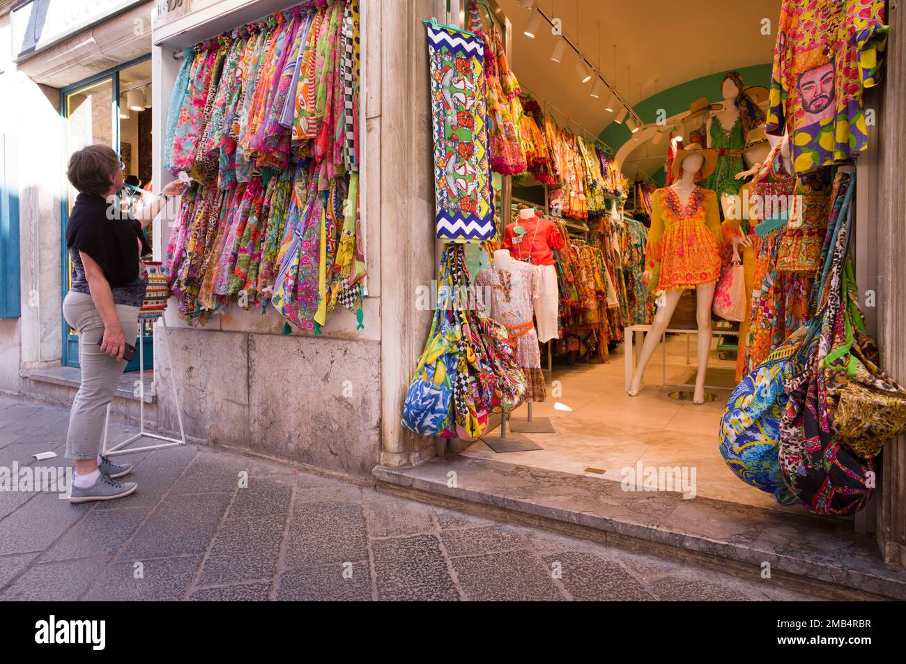 Ältere Frau, die typisch sizilianische Stoffe und Kleider ansieht und untersucht, Corso Umberto, Taormina, Sizilien, Italien Stockfoto