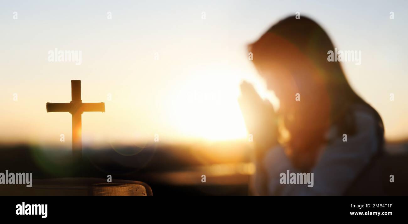 Der rote helle Hintergrund des Sonnenuntergangs, die Heilige Bibel, das Kreuz Jesu und ein christliches Gebet mit beiden Händen zusammen Stockfoto