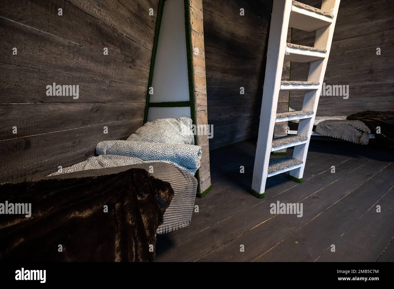 Einzelbetten mit sauberer Bettwäsche in Nischen aus Holz. Pyramidenförmiges Bungalow Sommerhaus. Abgewinkelte umgebene Wände, weiße Treppen, grauer horizontaler Arbeitsplatz Stockfoto