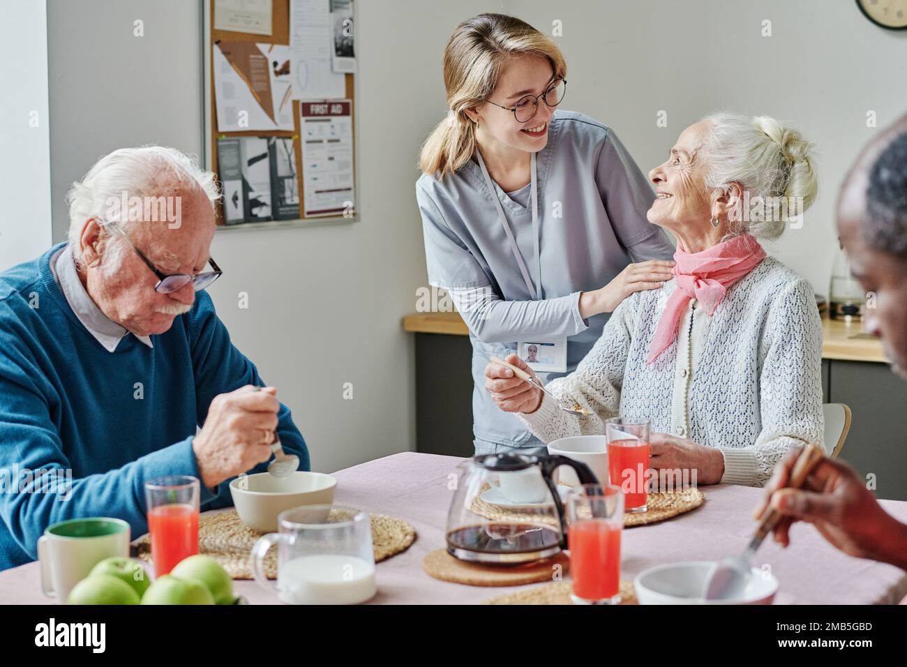 Betreuer spricht mit Senioren und hilft ihnen, während sie am Tisch essen Stockfoto