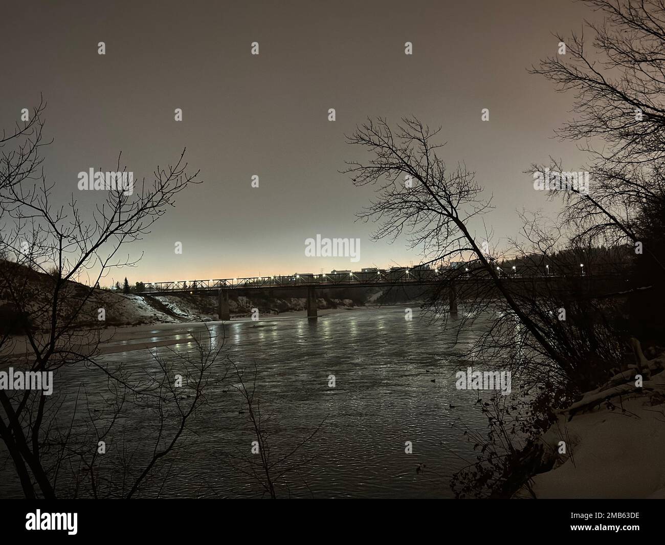 Eine Brücke über den Fluss, die bei Sonnenaufgang von der verschneiten Küste eingefangen wurde Stockfoto