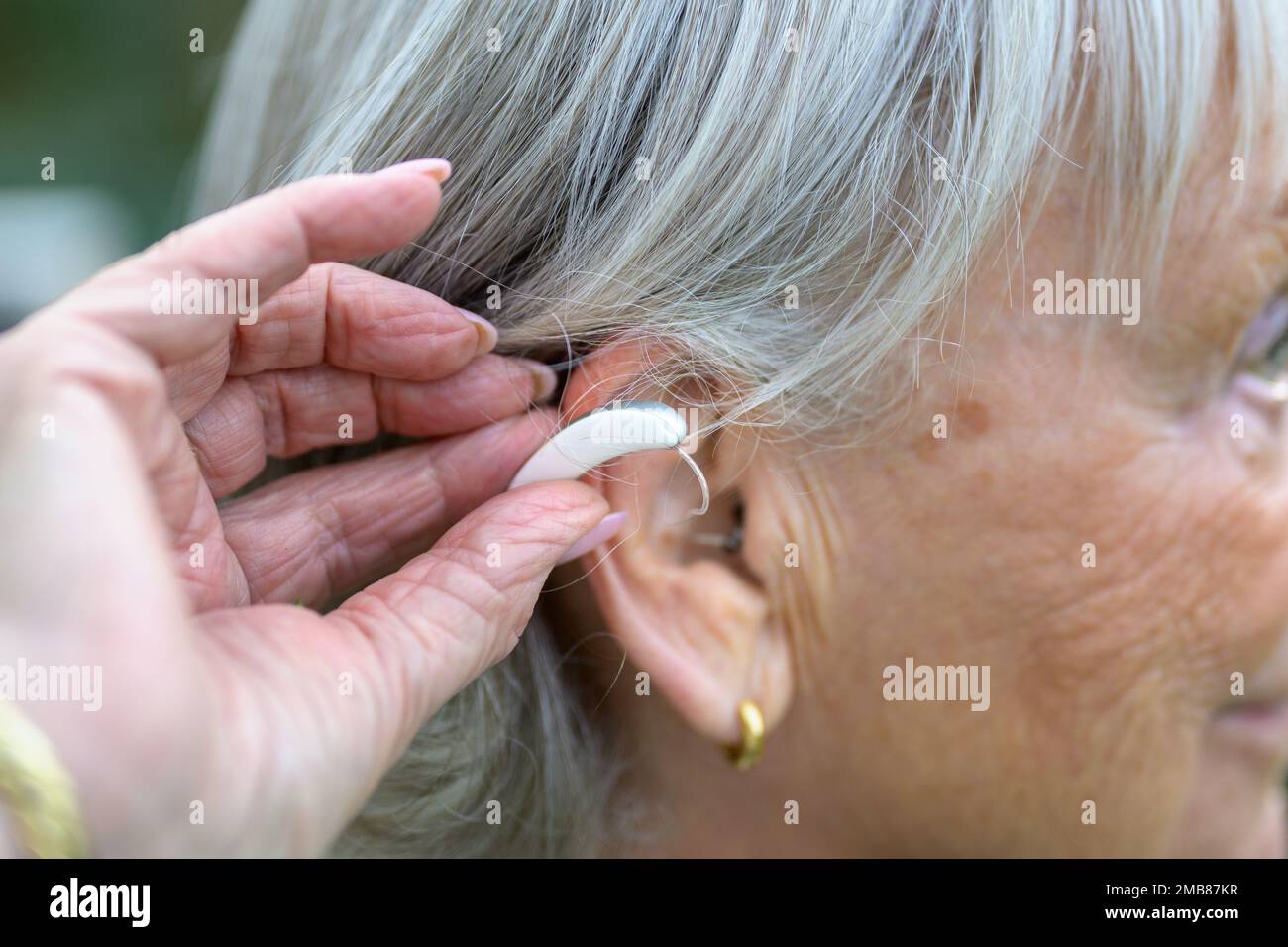 Ältere, grauhaarige, taube Dame in den 80ern, mit einem Hörgerät im Ohr, das sie gerade reingesteckt hat Stockfoto