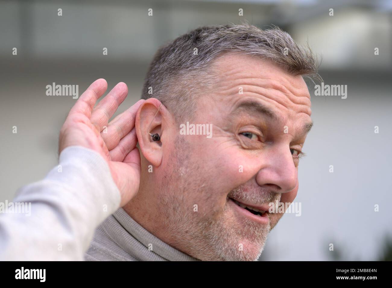 Ein Mann mit grauhaarigen Schwerhörigen im mittleren Alter zwischen 50 und 60 Jahren hält seine Hand hinter dem Ohr, in dem er ein Hörgerät hat Stockfoto