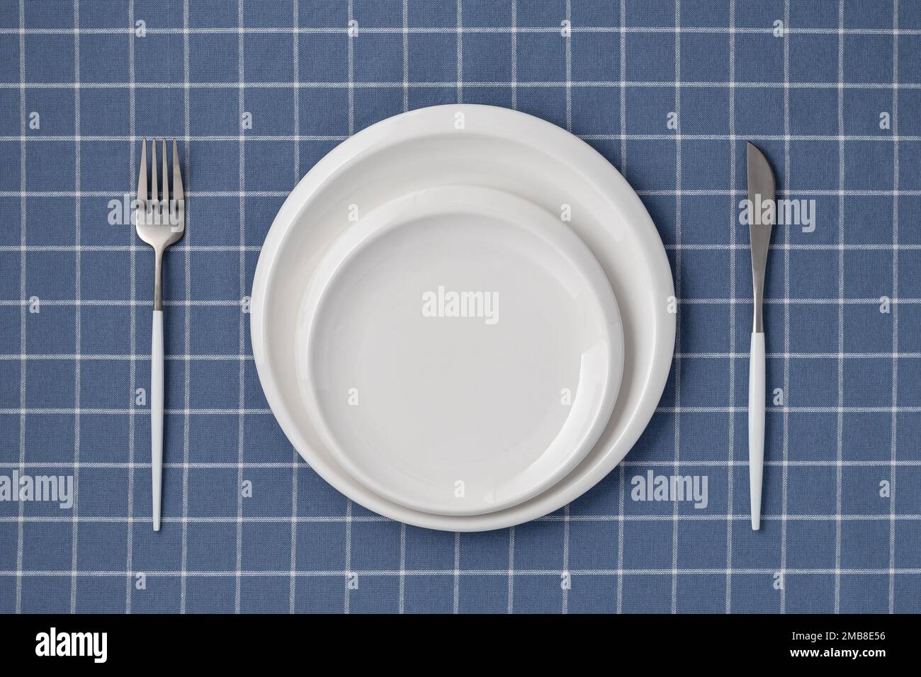 Weißer leerer Teller und Besteck auf einem blauen Tischtuch mit Karomuster, Blick von oben. Flach liegendes Design. Keramikutensilien, Servieren. Tablewar Stockfoto