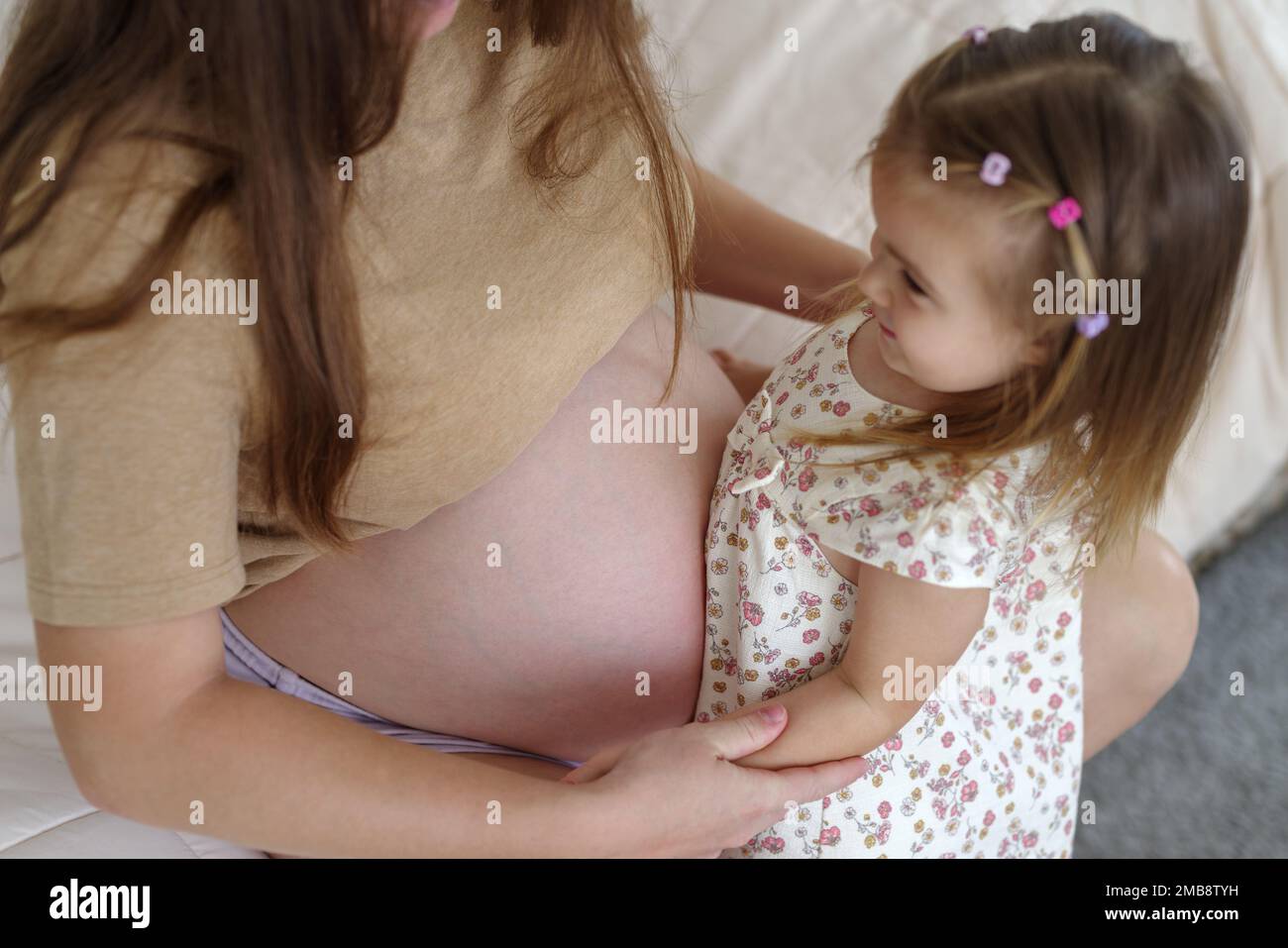 Eine schwangere weiße Mutter und ihre Tochter sind im Wohnzimmer. Die Tochter berührt den Bauch ihrer Mutter Stockfoto