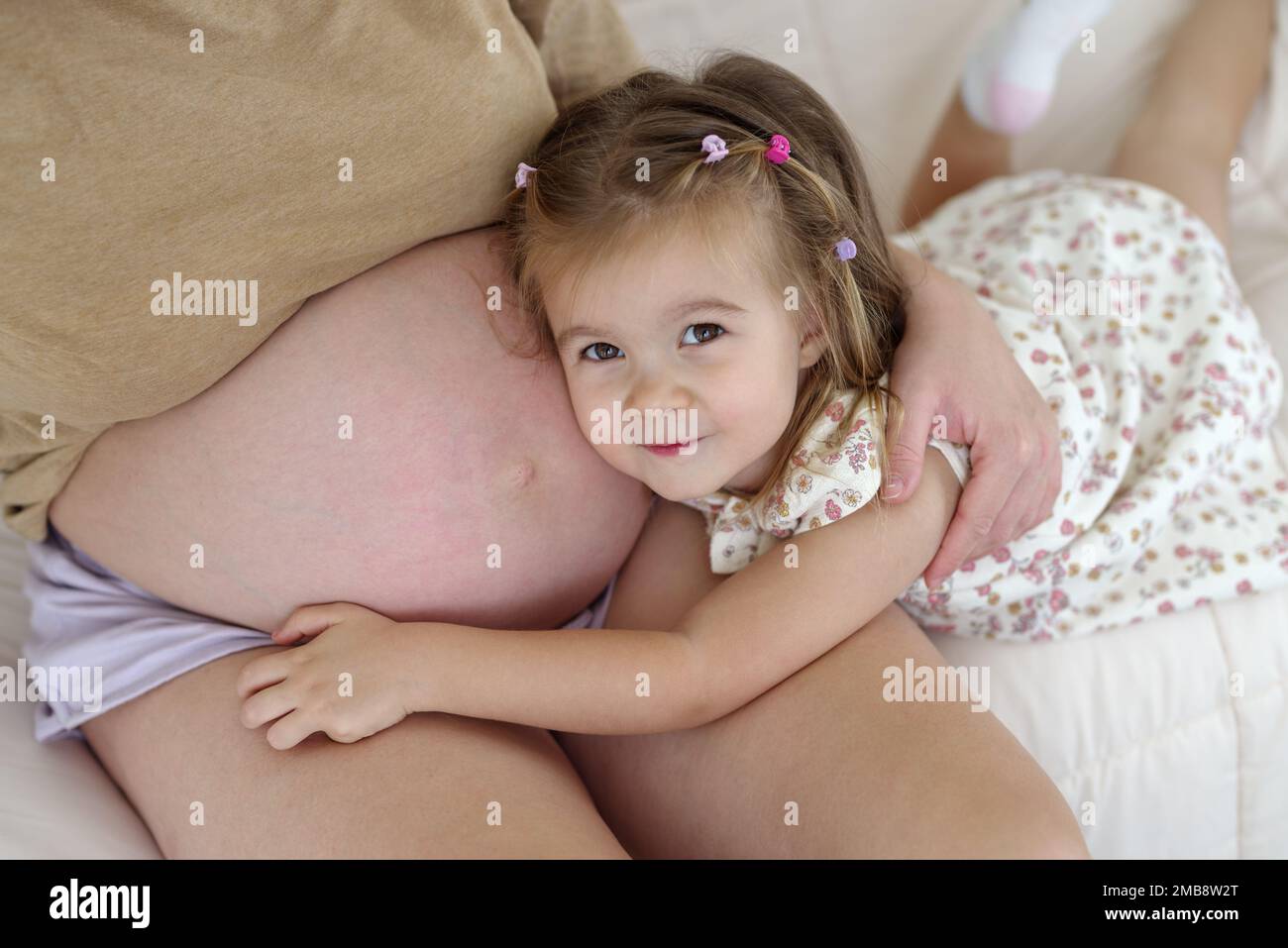 Eine schwangere weiße Mutter und ihre Tochter sind im Wohnzimmer. Die Tochter berührt den Bauch ihrer Mutter Stockfoto