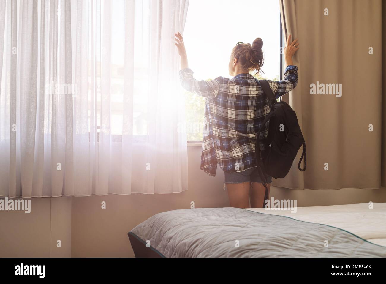 Eine Touristin mit Rucksack, die im Hotelzimmer steht, zieht die Vorhänge zurück und schaut aus dem Fenster. Stockfoto