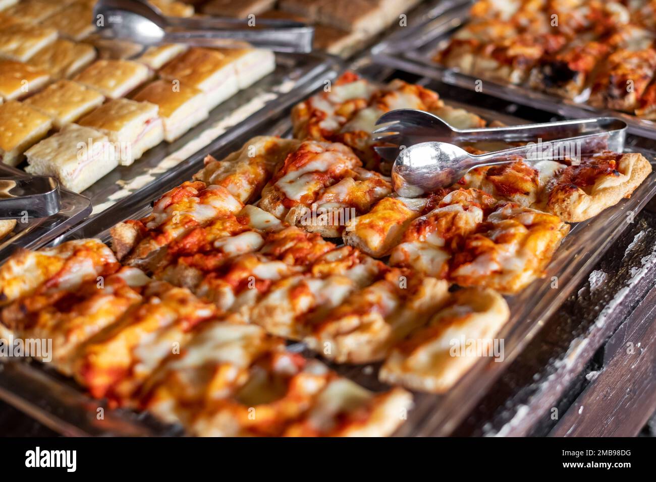 Von oben auf schmackhafte gebackene Pizzastücke, die auf ein Tablett gestellt wurden, mit einer Zange auf einem Selbstbedienungstisch Stockfoto