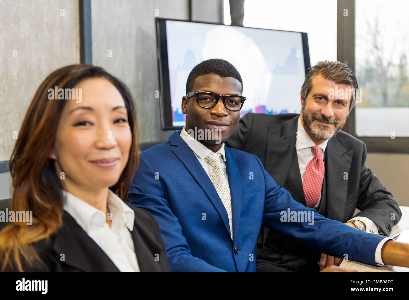 Gruppe positiver und vielfältiger Mitarbeiter in formeller Kleidung, die zusammen am Tisch sitzen und in einem modernen Büro in die Kamera schauen Stockfoto
