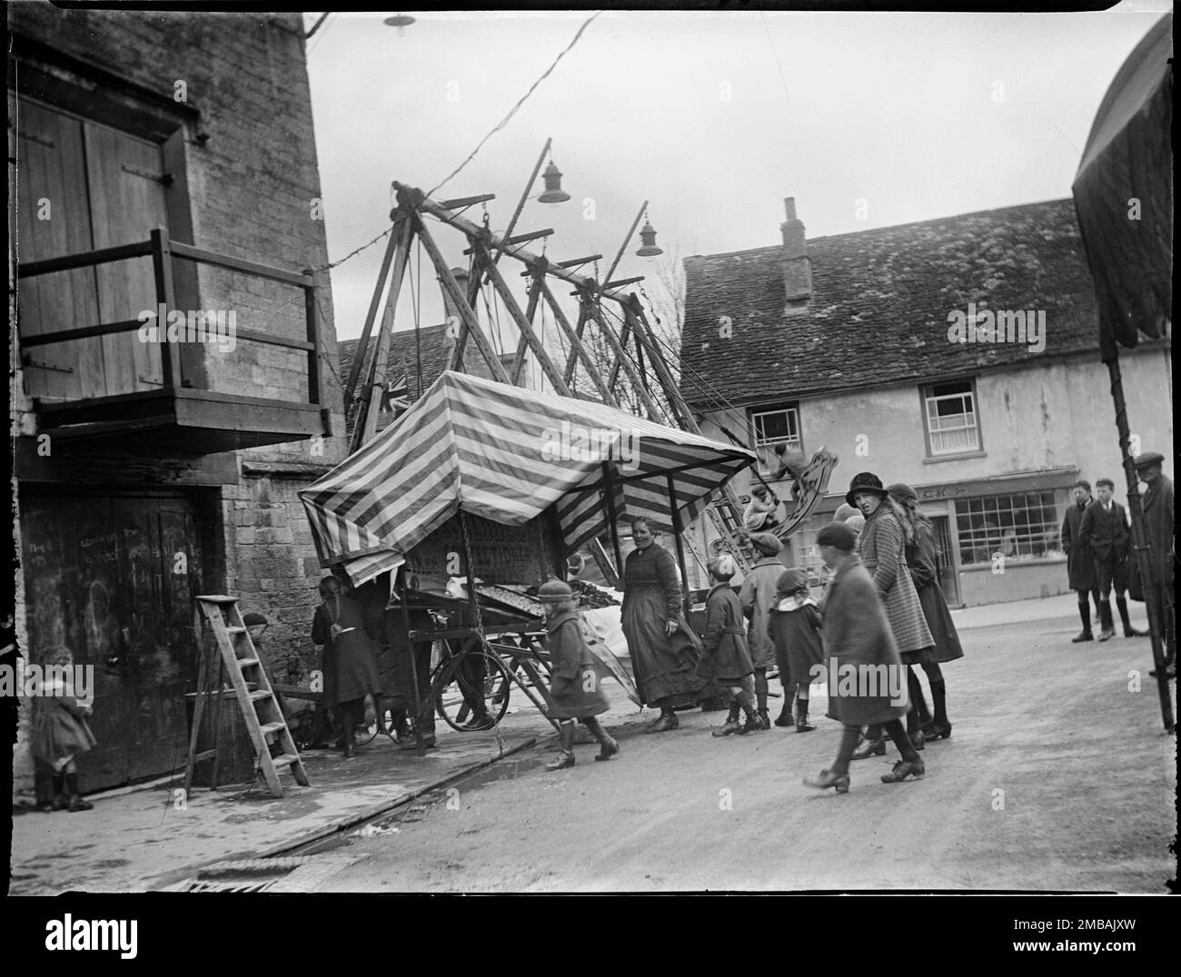 High Street, Burford, West Oxfordshire, Oxfordshire, 1924. Ein Blick auf den Jahrmarkt in Burford von der Kreuzung von Priory Lane mit der High Street aus, wobei Kinder neben einem Stand stehen und eine Fahrt mit dem Schaukelboot im Hintergrund stattfindet. Stockfoto