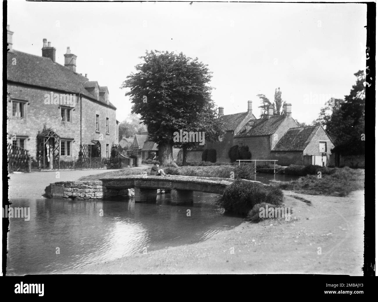 Lower Slaughter, Cotswold, Gloucestershire, 1928. Ein Blick nach Osten entlang des River Eye, wobei ein kleiner Junge auf einer Klapperbrücke sitzt, die auch als flussaufwärts Brücke bekannt ist, mit Häusern dahinter. Stockfoto