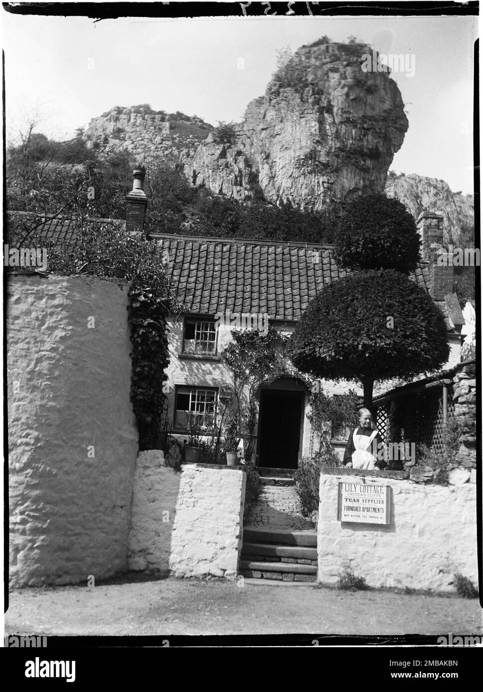Lily Cottage, Cliff Road, Cheddar, Sedgemoor, Somerset, 1907. Mrs. Main steht im Vorgarten von Lily Cottage mit dem Haus hinter ihr und Lion Rock auf dem Hügel darüber. Im Negativindex für die Sammlung bezeichnet der Fotograf die Frau, die auf dem Foto als Frau Main dargestellt wird. Das ist wahrscheinlich Mrs. Charlotte Main, die Erfrischungsräume leitete, die im Kelly's Directory von Somerset, 1902, aufgeführt sind. Stockfoto