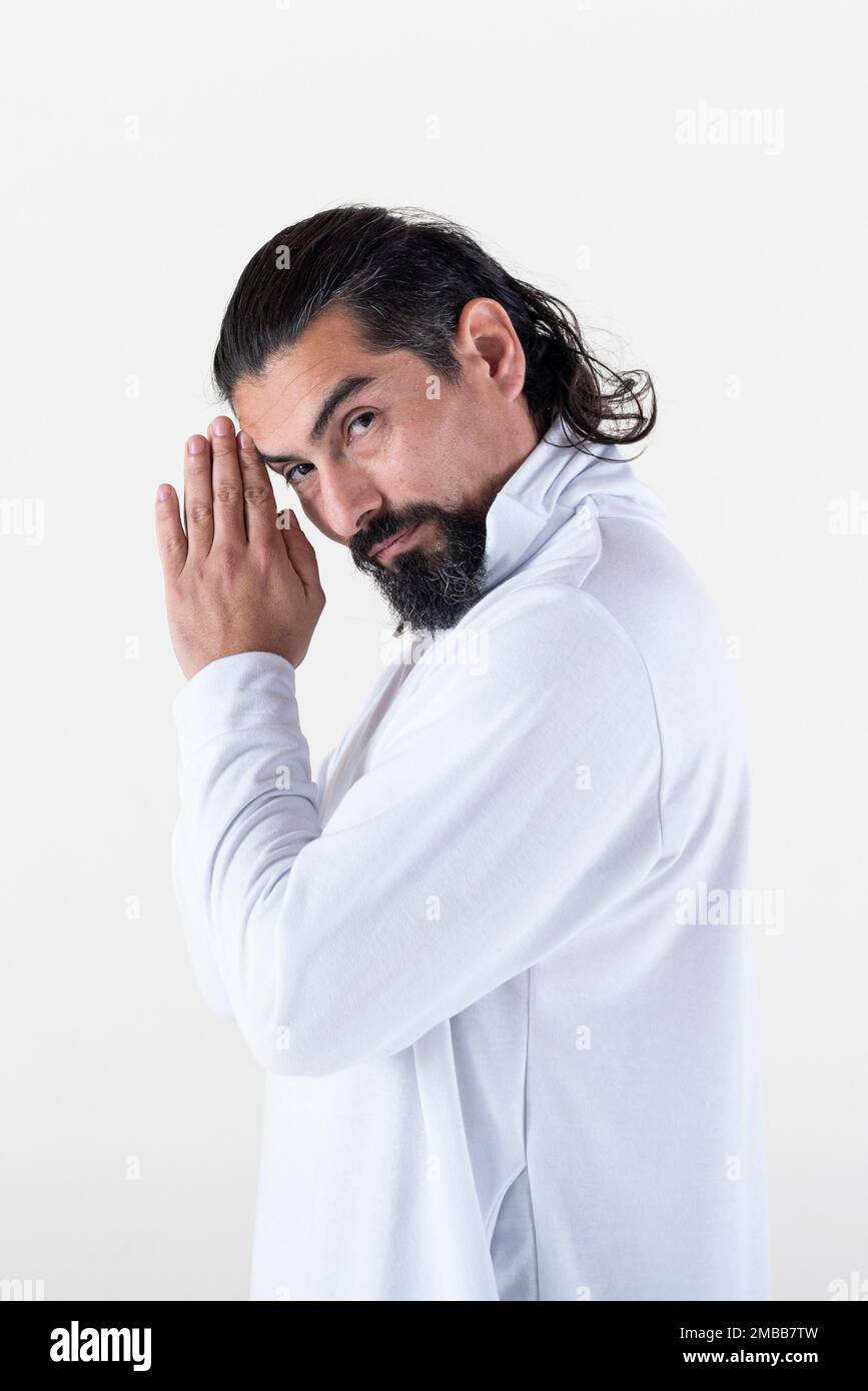 Ein Mann, der weiß gekleidet ist und Hände umschließt, während er über weißen Hintergrund in die Kamera schaut. Tadasana Yoga-Pose. Studioaufnahme. Stockfoto