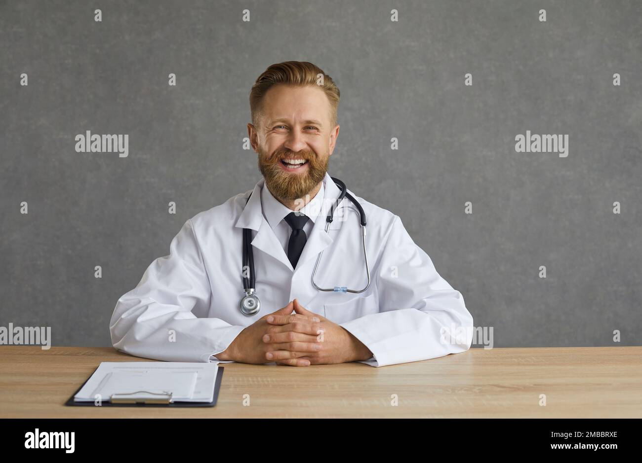 Erwachsener, weißer, männlicher Arzt, der einen medizinischen Mantel trägt und lacht am Schreibtisch Stockfoto