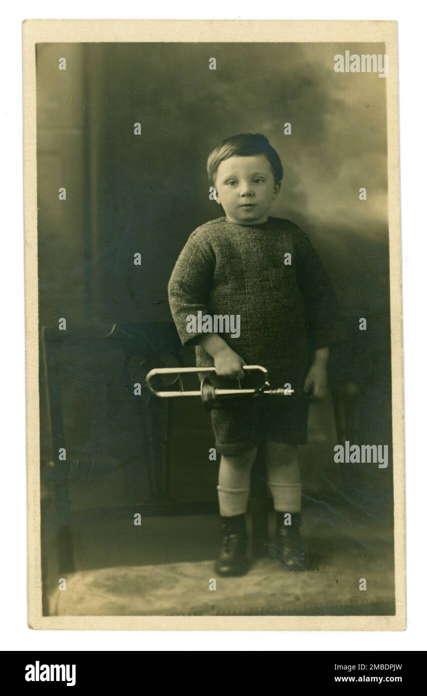 Originale, charmante Postkarte aus den frühen 1900er Jahren mit einem niedlichen kleinen Jungen mit hausgemachter TiN-Spielzeugtrombone, Musikinstrument, vielleicht einem Studio-Requisit oder Lieblingsspielzeug. Der süße kleine Junge trägt Schnürstiefel, einen handgestrickten Pullover und dazu passende Shorts. Etwa 1910, 1920 in Großbritannien Stockfoto