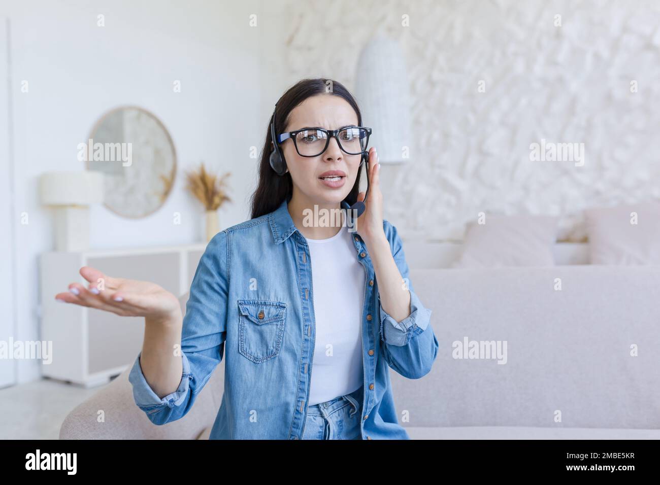 Besorgte junge Frau sitzt zu Hause auf dem Sofa im Headset, spricht über Mikrofon, Online-Beratung. Sie schaut in die Kamera, erklärt, Gesten mit seinen Händen. Stockfoto