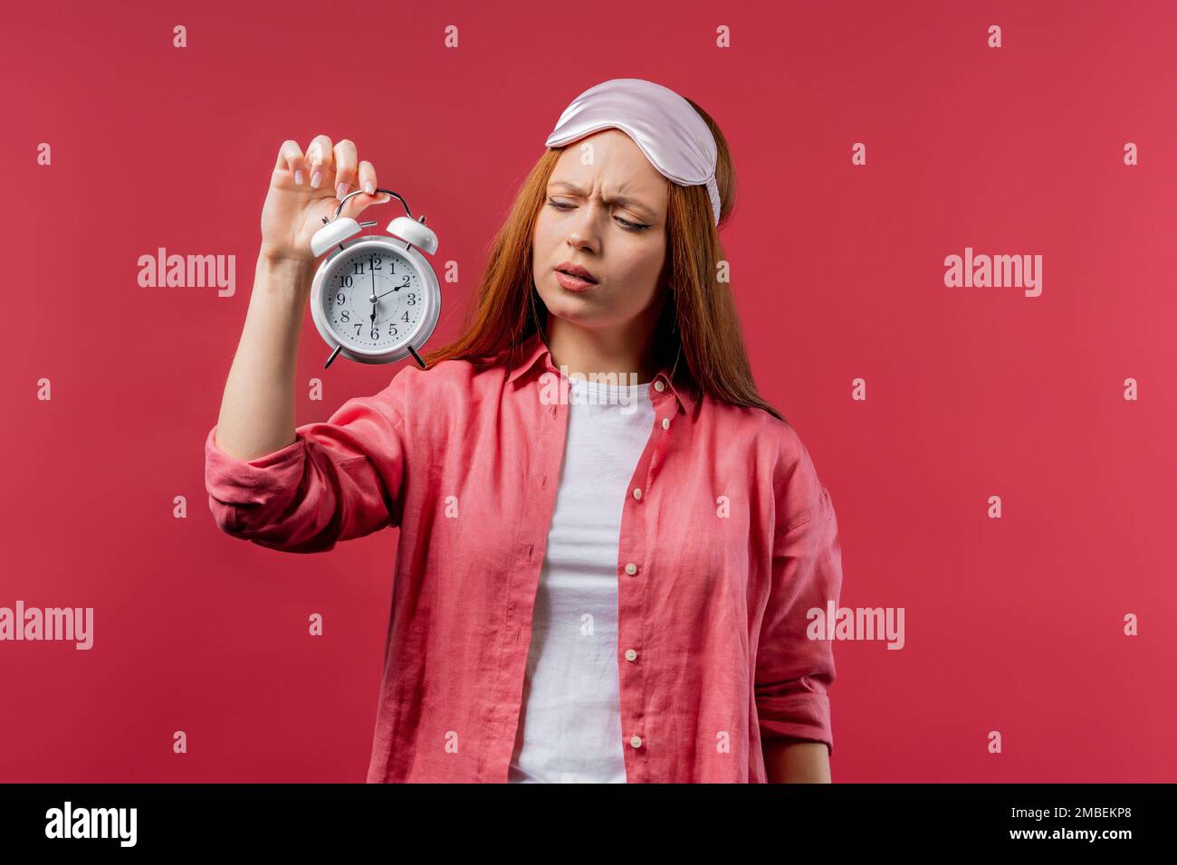 Wachte auf von Wecker, schläfrige Frau, die es in der Hand hielt. Rosafarbener Hintergrund. Früh um 6 Uhr morgens. Die faule Dame hat nicht genug Schlaf bekommen, Konzept von Stockfoto