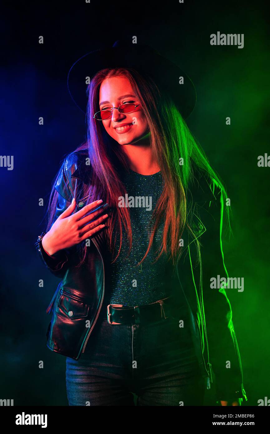 Modische Frau in mehrfarbigem Neonlicht. Rauchiger Hintergrund, Nachtparty, Disco-Club. Z-Generation rätselhafte schöne Dame in Lederjacke, Hut Stockfoto