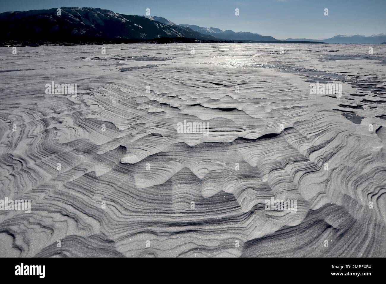 Texturen und Linien im Schneefall auf dem gefrorenen See mit blauem Himmel und bewaldeten Bergen im Hintergrund. An einem dieser knackigen arktischen Wintermorgen mit Stockfoto
