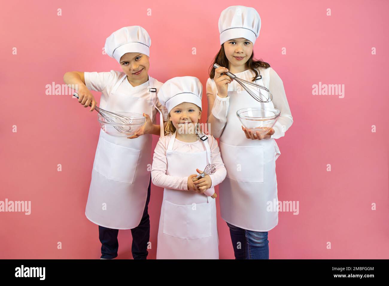 Fröhliche Kinder mit Kappen und Schürze, die Schneebesen und Tassen auf pinkfarbenem Hintergrund halten Stockfoto
