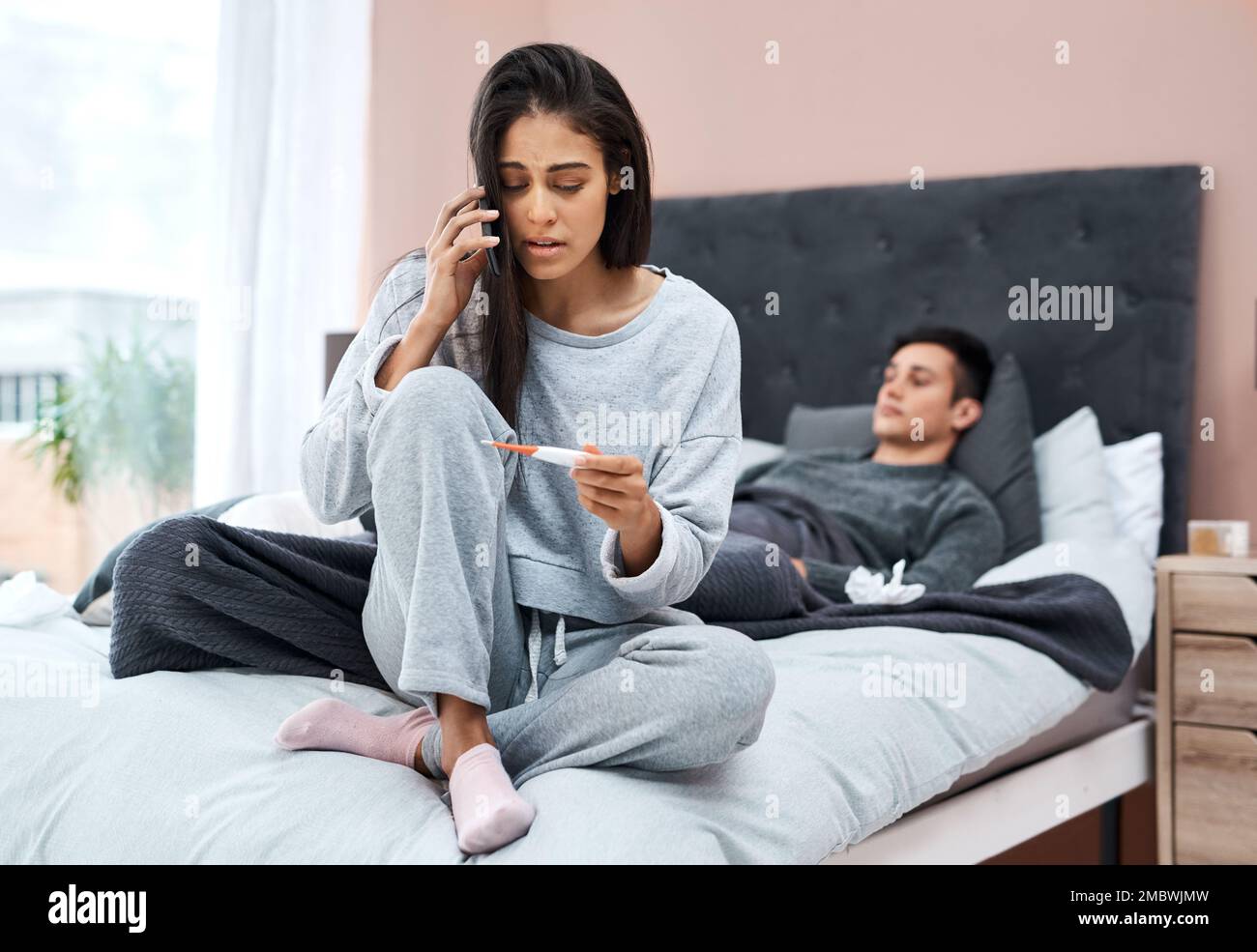 Die Hotline für Gesundheit. Eine junge Frau, die mit ihrem Mann zu Hause ein Smartphone und ein Thermometer benutzt, während sie sich von einer Krankheit erholt. Stockfoto