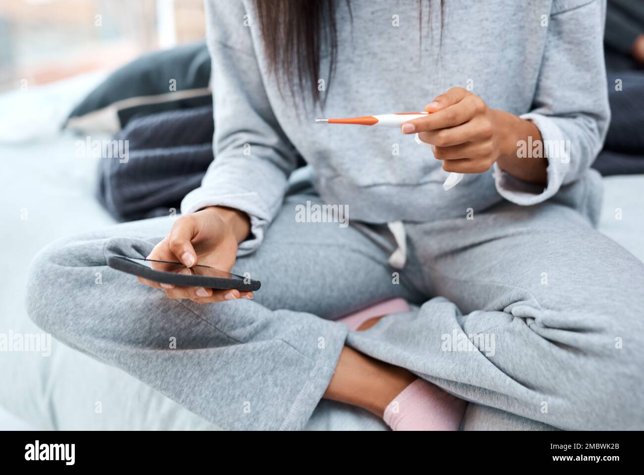 Medizinische Versorgung wird mobil. Eine nicht wiedererkennbare Frau, die ein Smartphone und ein Thermometer benutzt, während sie sich von einer Krankheit zu Hause erholt hat. Stockfoto