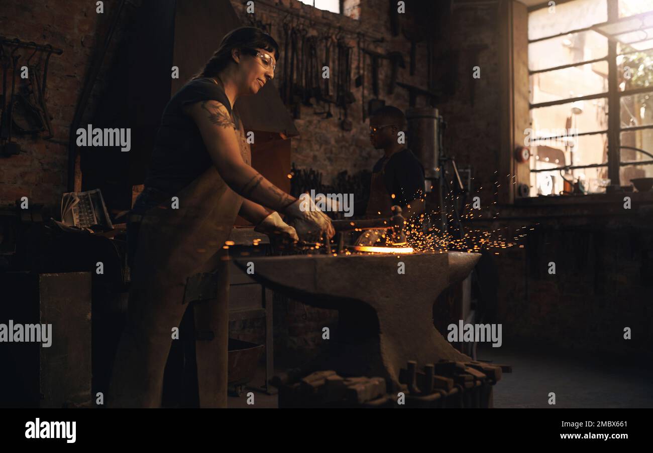 Die Stahlwelt hat viel zu verdanken für ihre Arbeit. Eine junge Frau, die in einer Gießerei mit einem Hammer auf einen heißen Metallstab schlägt. Stockfoto