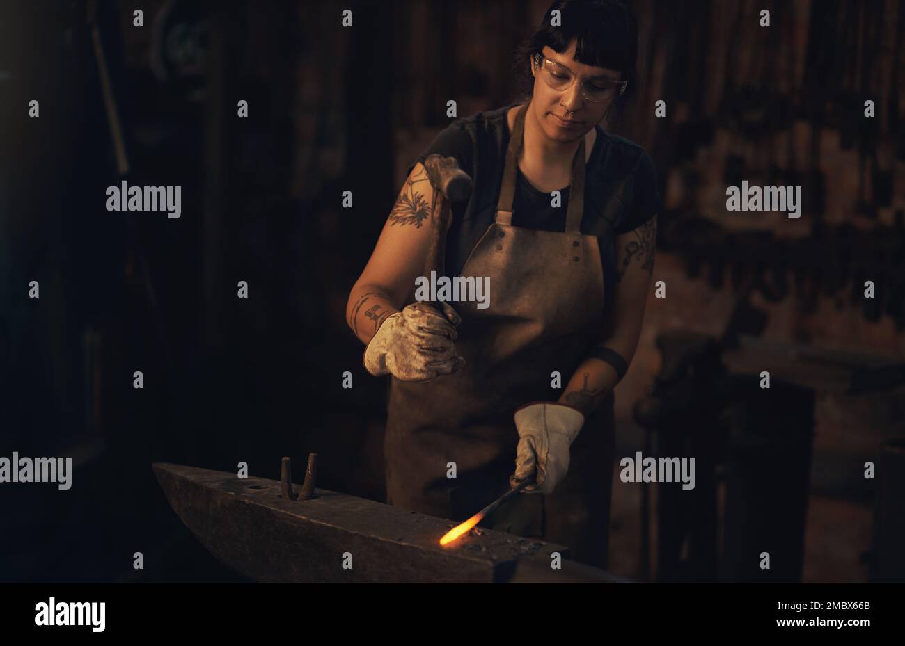 Das heißeste Talent in der Metallwerkindustrie. Eine junge Frau, die in einer Gießerei mit einem Hammer auf einen heißen Metallstab schlägt. Stockfoto