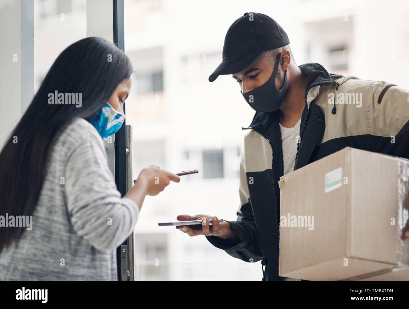 Eine kontaktlose Lieferung sorgt für Sicherheit. Ein maskierter junger Mann und eine maskierte junge Frau, die Smartphones während einer Lieferung nach Hause benutzen. Stockfoto