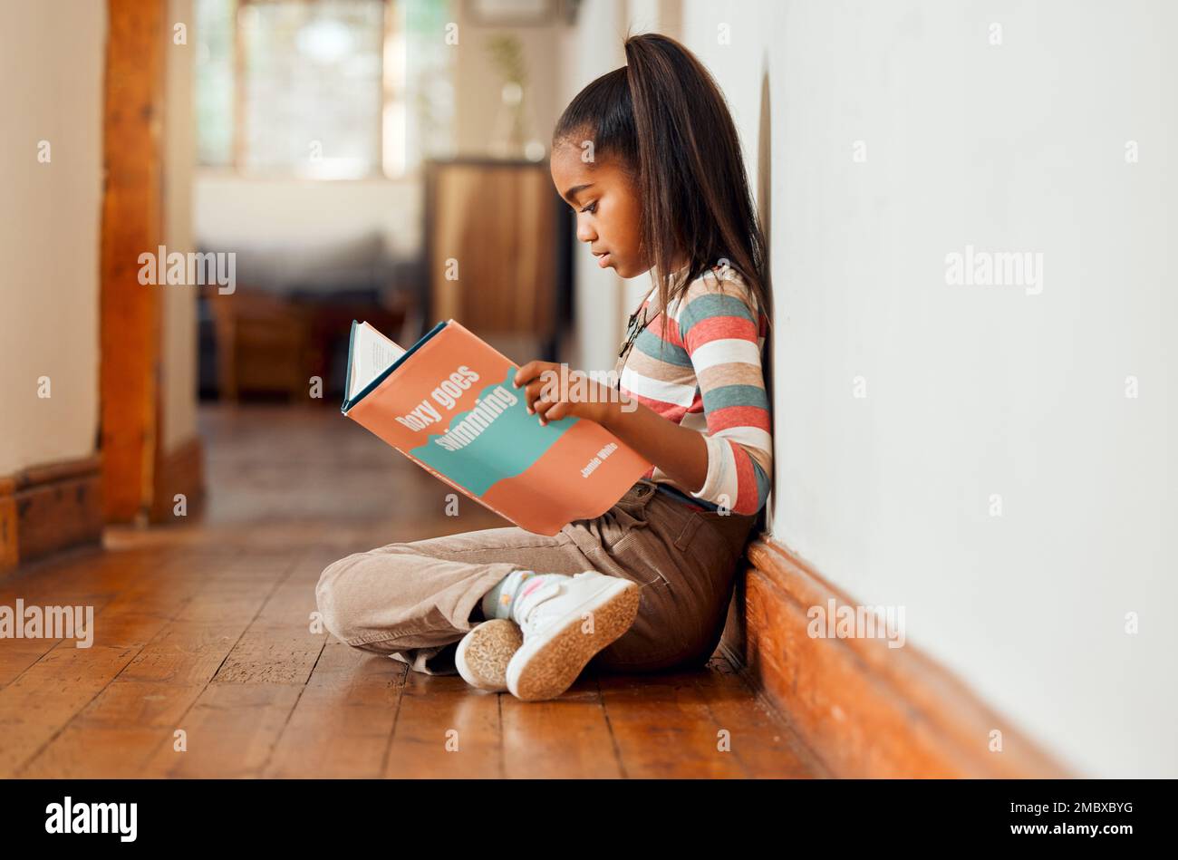 Kleines Mädchen, Buch und lies auf Holzboden, um Geschichten zu erzählen, zu lernen oder Bildung zu lernen, entspannt zu Hause mit einem Lächeln. Glückliches Kind, das lächelt, sitzt und Stockfoto