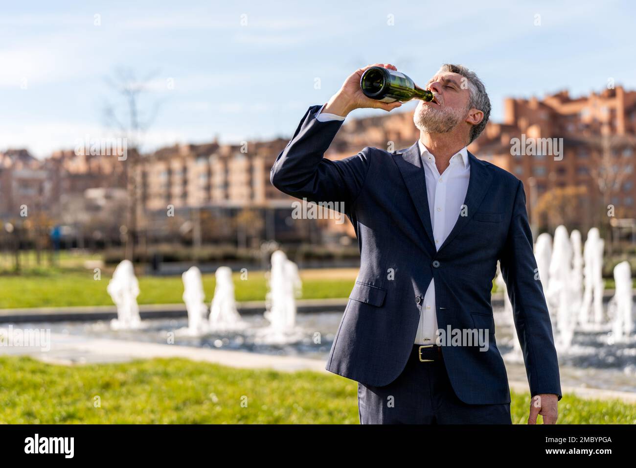Geschäftsmann mittleren Alters mit Bart im formellen Anzug, der Champagner aus einer Flasche trinkt, während er auf der Straße gegen den Brunnen steht Stockfoto