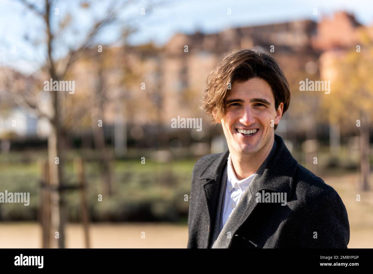 Fröhlicher junger Geschäftsmann in stilvollen Outfits, der lächelt und vor die Kamera schaut, während er Zeit im Park verbringt Stockfoto