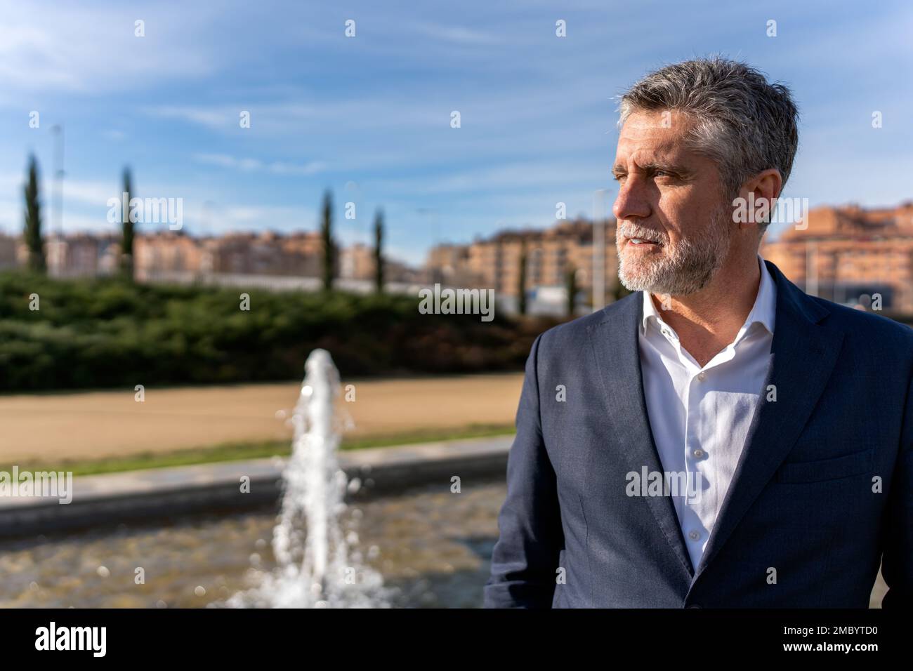Ein durchdachter älterer Geschäftsmann mit grauen Haaren, der einen formellen Anzug trägt, steht neben dem Brunnen und blickt weg, während er die atemberaubende Landschaft bewundert Stockfoto