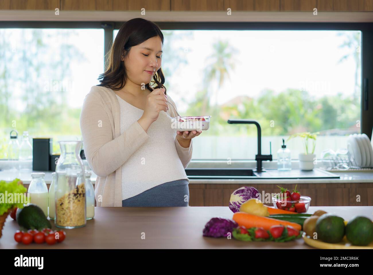 Eine wunderschöne asiatische schwangere Frau, die beim Frühstück zu Hause in der Küche Müsli isst und das Essen genießt. Leckeres Essen für Schwangere Stockfoto
