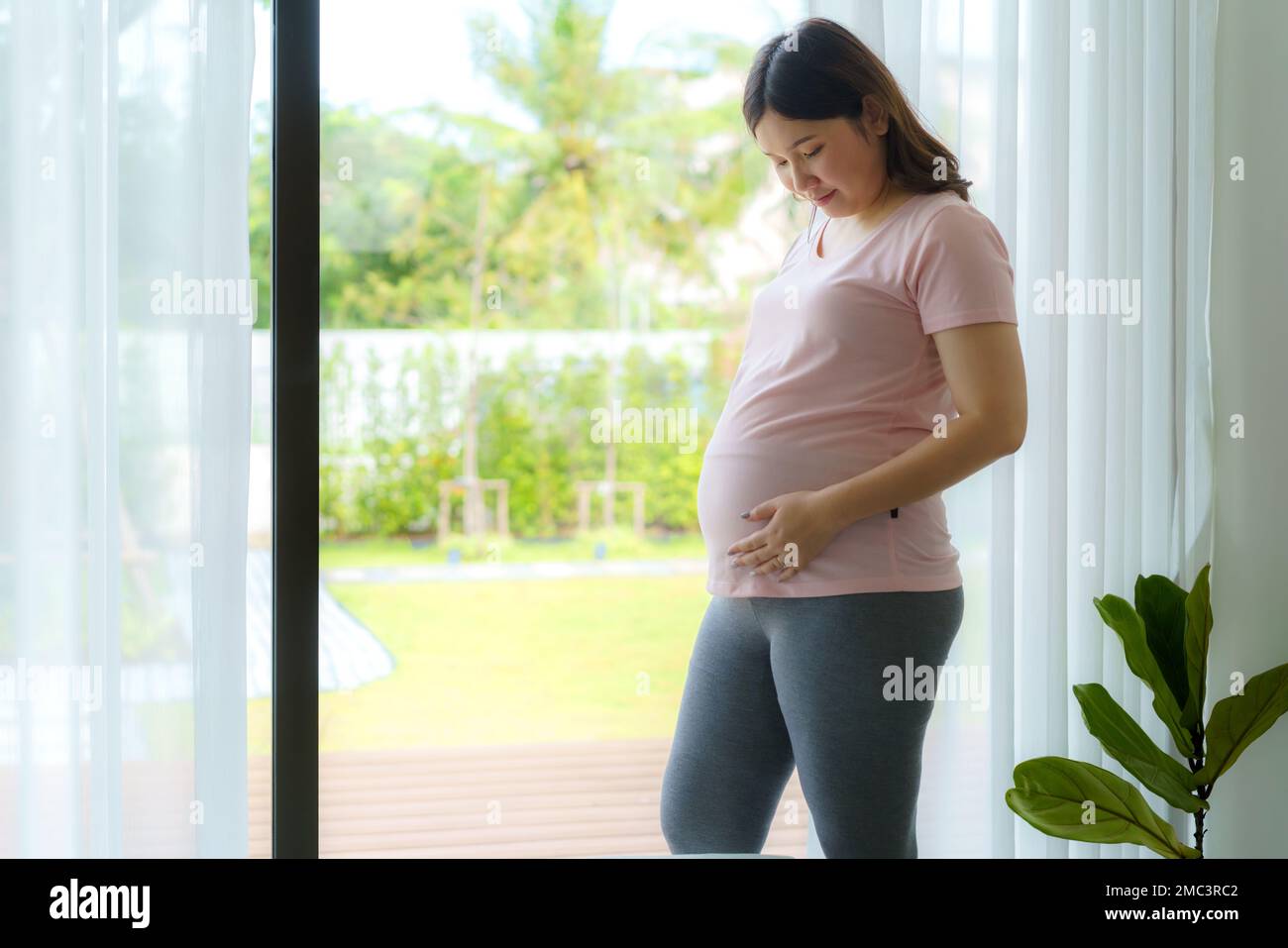 Schwangerschaft, Menschen Ruhe und Erwartung Konzept - Asiatische, lächelnde, glückliche schwangere Frau, die am Fenster steht und ihren Bauch zu Hause berührt. Stockfoto