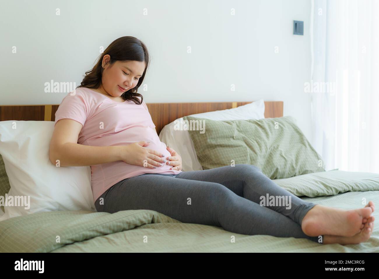 Schwangerschaft, Ruhezustand und Erwartungskonzept - Asiatische, lächelnde, glückliche, schwangere Frau, die im Bett sitzt und zu Hause ihren Bauch berührt. Stockfoto