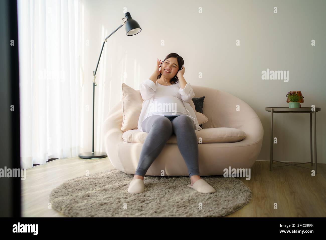 Porträt einer schwangeren Happy Asian Woman, die zu Hause auf dem Sofa sitzt und ein Smartphone benutzt, um Musik über Kopfhörer zu hören. Stockfoto