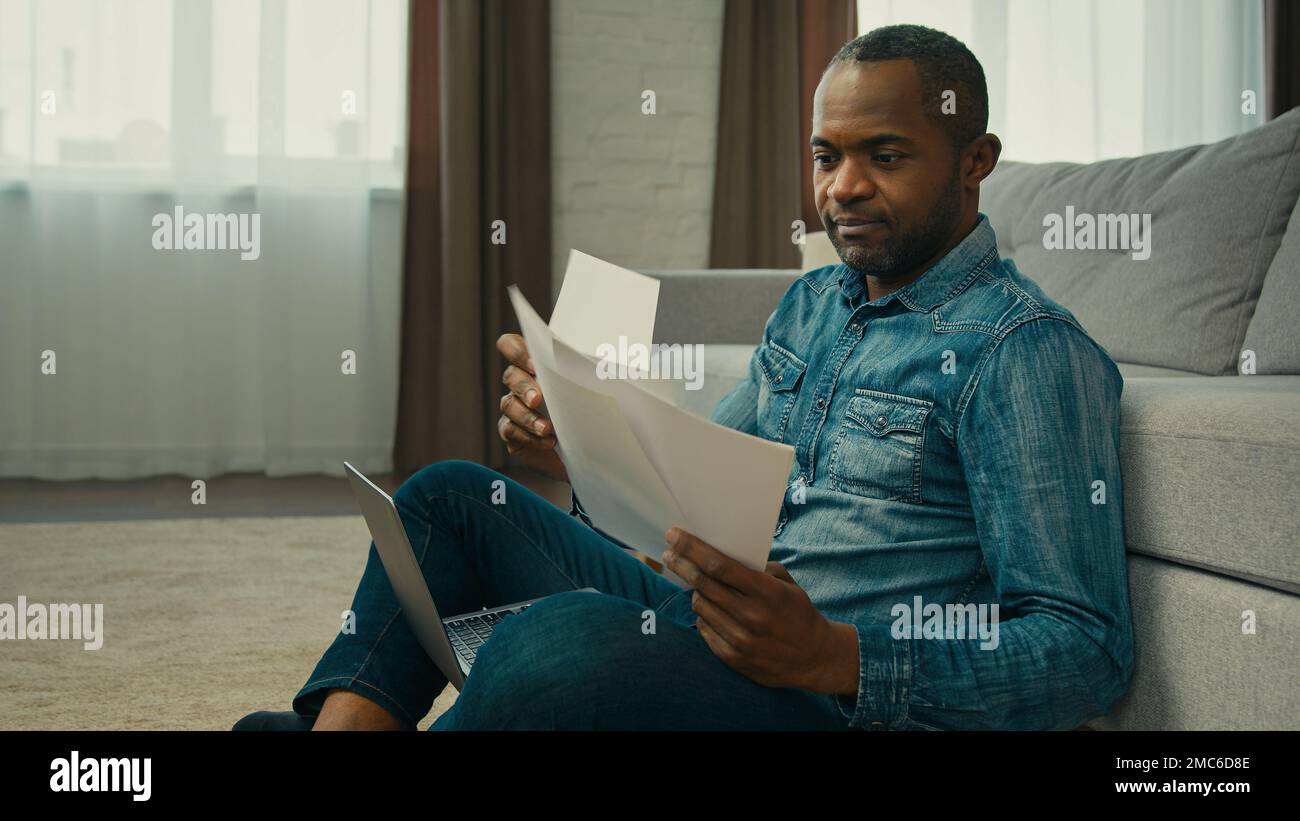 Ein reifer afroamerikanischer Mann, der im Wohnzimmer sitzt und Dokumente liest, ein ethnischer Geschäftsmann, der mit Zeitungen arbeitet, analysiert die Finanzprüfung Stockfoto