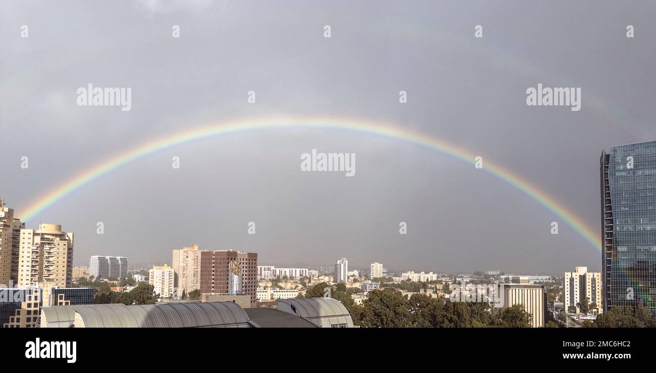 Ein wunderschöner Regenbogen springt aus einem wolkigen Himmel über einer großen Stadt mit einigen Hochhäusern vor einem wolkigen Himmel, bei dem die Sonne versucht, herauszukommen Stockfoto